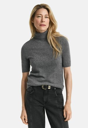Donna con lunghi capelli biondi che indossa un maglione grigio a maniche corte con colletto alto, infilato in jeans neri con una cintura nera.