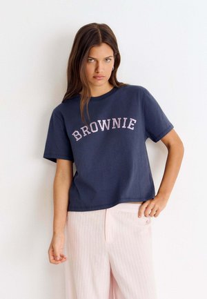 Jeune femme aux longs cheveux bruns portant un T-shirt bleu marine avec l'inscription "BROWNIE" et un pantalon rose pâle, debout devant un mur blanc uni.