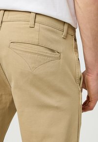 Beige Chinos mit glatter Textur, standardmäßiger Taille und einer Gesäßtasche mit spitzer Gestaltung. Der Stoff ist leicht und maßgeschneidert.