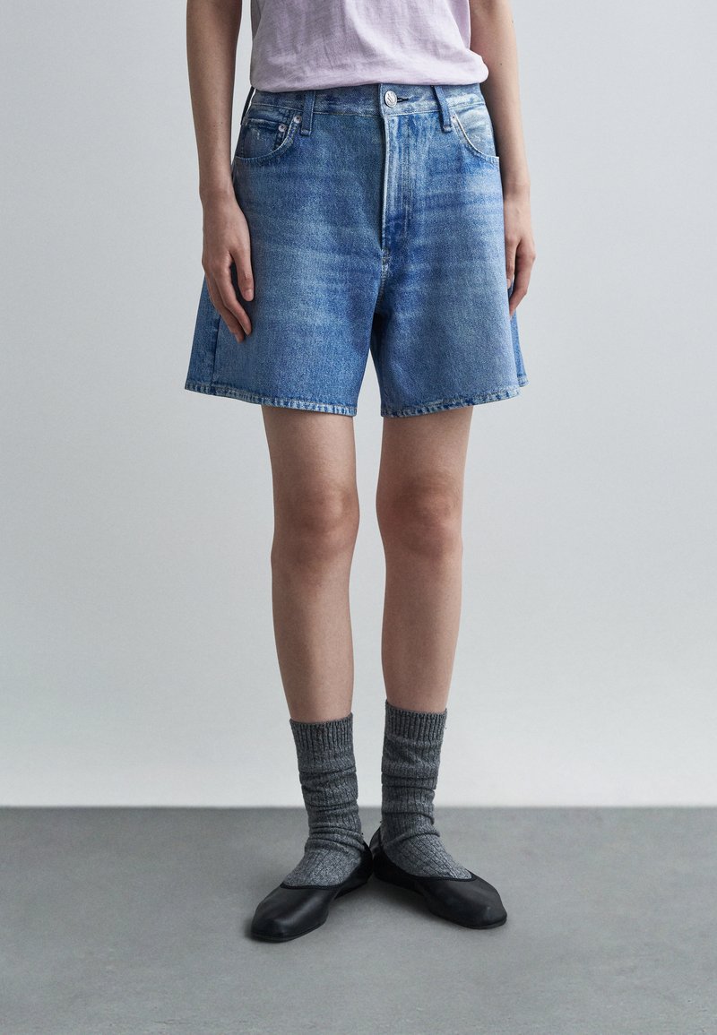 Osoba oblečená v modrých džínsových šortkách, šedých rebrovaných ponožkách, čiernych plochých topánkach a svetlofialovej košeli, stojaca na sivom podlaží s jednoduchým pozadím.