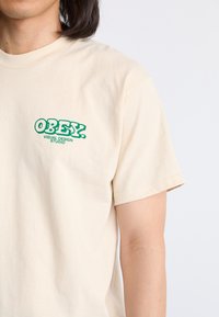 Camiseta de algodón beige con texto impreso en verde "OBEY. VISUAL DESIGN STUDIO" en el lado izquierdo del pecho. Mangas cortas y escote redondo.