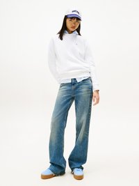 Sudadera blanca, camisa de botón ligera azul debajo, zapatillas deportivas azules claras y jeans de pierna ancha descoloridos. Gorra con el logo "TOMMY JEAN" en morado.