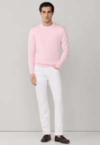 Uomo che indossa un maglione a maniche lunghe rosa chiaro, jeans bianchi e mocassini con nappine marrone scuro, in piedi contro uno sfondo semplice grigio chiaro.