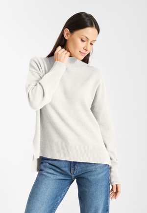 Lichtgrijze trui met hoge kraag, geribbelde manchetten en zoom. Het materiaal lijkt zacht; heeft zijsplitten. Gecombineerd met blauwe jeans.