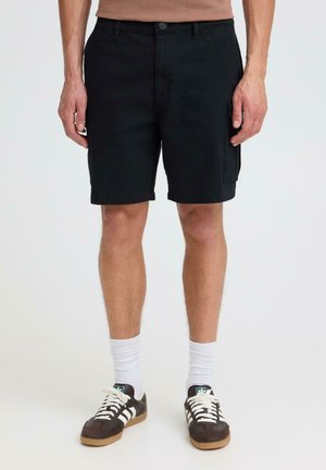 Man die zwarte cargoshorts, witte sokken en zwarte Adidas sneakers draagt, staand tegen een effen lichte achtergrond.
