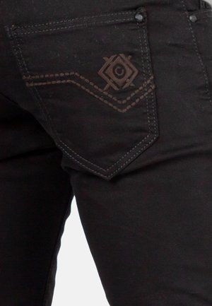 Schwarze Jeans aus Denim mit einer Gesäßtasche, die ein geprägtes Diamantmuster und Kontrastnähte aufweist. Glatte Textur mit metallischen Akzenten.