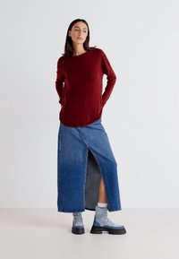 Bordeaux lange mouw top gecombineerd met een denim maxi rok met een split aan de voorkant. Model draagt grijze enkellaarsjes met zwarte zolen.