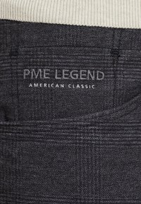 Tissu gris foncé avec un motif subtil. Présente "PME LEGEND" et "AMERICAN CLASSIC" brodés sur une poche arrière.