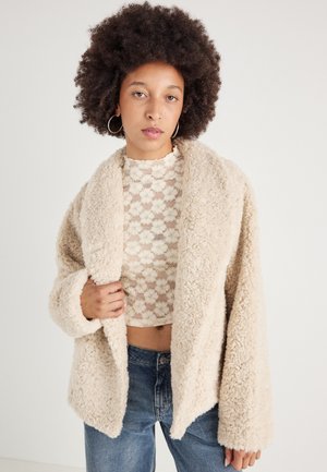 Giacca in fleece beige con una finitura morbida e testurizzata, davanti aperto e maniche larghe, abbinata a un top corto in pizzo floreale e jeans di denim.