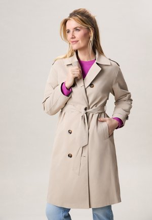 Vrouw met beige trenchcoat met ceintuur over paarse top en blauwe spijkerbroek, staand met één hand in haar zak tegen een effen achtergrond.