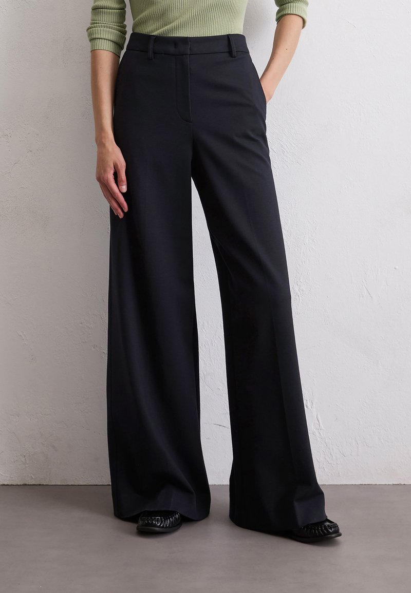 Schwarze weite Hose mit glatter Textur und tailliertem Design, mit flachem Bund, Seitentaschen und kombiniert mit schwarzen Schuhen.