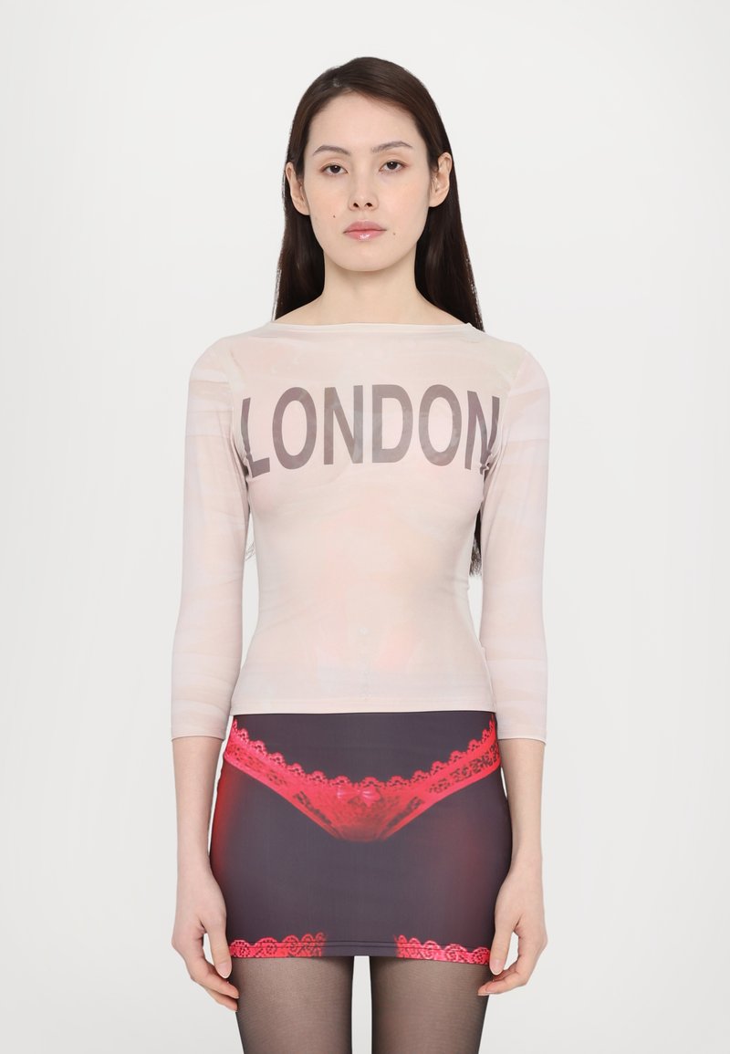 Transparente langärmelige Bluse mit "LONDON" in fettem schwarzen Text, kombiniert mit einem taillierten Rock, der ein grafisches Spitzenmuster in Rot auf dunklem Stoff zeigt.