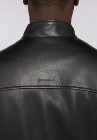 Černá kožená bunda s hladkým povrchem, s límcem ve tvaru pásku a s našitým logem "TRUSSARDI" na horní části zad.