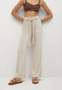Pantalon large beige en tissu léger, avec une taille nouée et une finition texturée, associé à des sandales à plateforme marron.