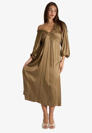 Donna che indossa un abito midi lucido verde oliva, con spalle scoperte, maniche a sbuffo e tacchi color crema con punta aperta, tenendo l'orlo dell'abito con una mano.