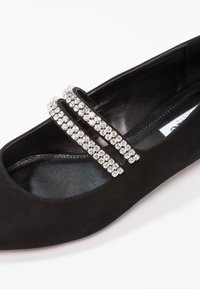 Zapato plano de ante negro con dos filas de brillantes strass plateados en la parte delantera. Presenta una puntera redondeada y un forro interior suave.