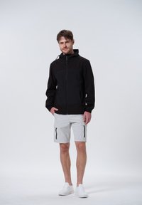 Chaqueta negra con cremallera y capucha, con un panel frontal liso y puños elásticos. Combinada con pantalones cortos cargo de color gris claro y zapatillas blancas.
