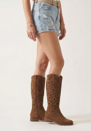 Kobieta w podniszczonych szortach z denimu i brązowych zamszowych kozakach za kolano z wyciętymi wzorami, stojąca na jednolitym tle.