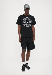 Svart t-skjorte med hvit "Versace Jeans Couture" logo, sammen med svarte shorts og svarte joggesko. Grå hettecap med matchende logo synlig.