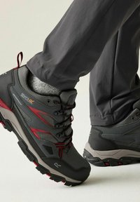 Persona con zapatos de senderismo impermeables grises y rojos con suelas negras y grises y pantalones gris oscuro.