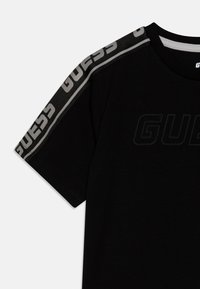 Svart T-skjorte laget av bomull, med 'GUESS'-logoen i teksturert trykk og et grått mønster på skulderen. Rund hals.