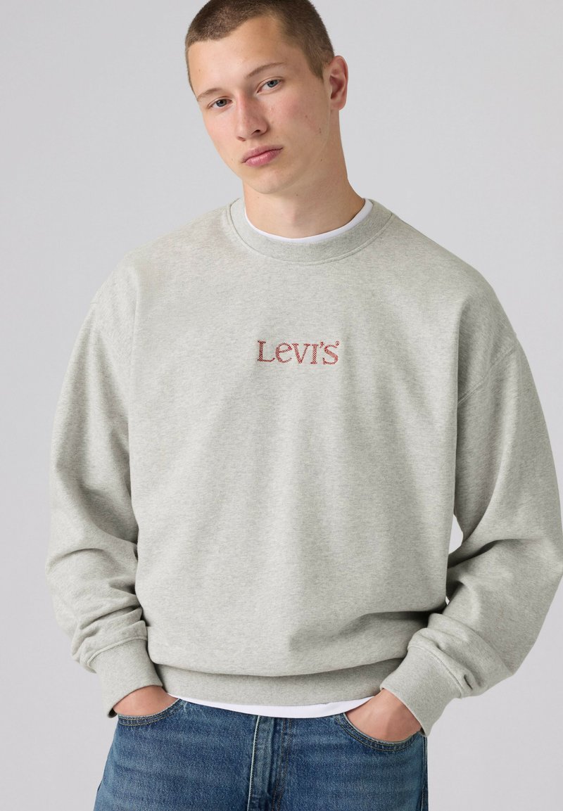 Giovane uomo con capelli corti che indossa una felpa Levi's grigio chiaro e jeans blu, in piedi con le mani nelle tasche davanti a uno sfondo semplice.