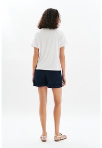 Top corto blanco de manga corta combinado con pantalones cortos azul marino, que cuentan con puños enrollados y bolsillos traseros. El modelo lleva sandalias color canela, de pie frente a un fondo liso.