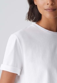 T-shirt en coton blanc avec des manches roulées, texture douce et col rond classique, partiellement visible sur l'épaule et le cou du modèle.