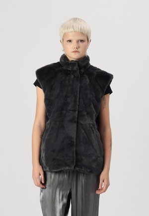 Svart faux fur-vest med høy krage, korte ermer, myk tekstur og frontklips. Båret over en svart skjorte og sølvfargede bukser.