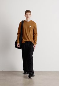 Καφέ μακρυμάνικη ριγέ μπλούζα με μικρό λογότυπο, σε συνδυασμό με μαύρες φαρδιές παντελόνες και μαύρα παπούτσια, κρατώντας ένα μαύρο τσαντάκι τύπου messenger.