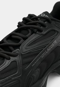 Μαύρο αθλητικό παπούτσι με υφή από συνθετικό υλικό, στρογγυλή μύτη, μαύρες κορδόνες και λογότυπο "LACOSTE" στο πλάι.