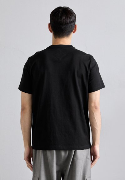 T-shirt en coton noir à manches courtes, col rond simple, et un petit motif en losange au dos. Porté avec un pantalon gris.
