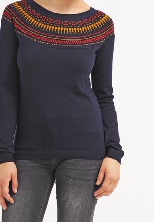 Femme portant un pull marine avec un empiècement à motifs rouges et orange et un jean gris, debout devant un fond blanc.