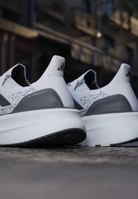 Paire de chaussures de course Adidas Ultraboost blanches et grises avec tissu tricoté moucheté, vues de dos sur une surface pavée en extérieur.
