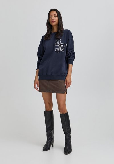 Námornícky sveter s predným logom, voľný strih a rebrované manžety, spárovaný s hnedou džínsovou sukňou a čiernymi vysokými čižmami.