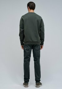 Felpa verde scuro con dettagli a coste sulle spalle, abbinata a jeans scuri e scarpe grigie, vista da dietro su uno sfondo chiaro.