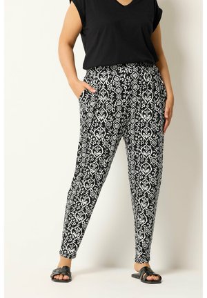 Personne portant un pantalon fuselé à motif noir et blanc, un haut noir à manches courtes et des sandales noires ouvertes à enfiler, debout avec une main dans la poche.