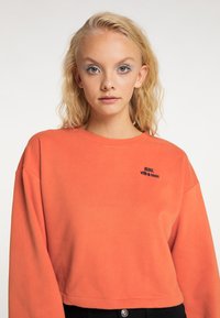 Sweatshirt scurt de culoare portocalie, cu guler rotund și mâneci lungi, având text brodat "REBEL cu un scop" în albastru închis.