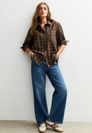 Brown plaid button-up shirt with rolled sleeves, paired with wide-leg blue jeans and white sneakers accented with brown stripes.