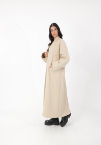 Cappotto lungo beige con una vestibilità rilassata, realizzato in materiale morbido, con maniche larghe e tasche. Abbinato a stivali platform neri in stile chunky.
