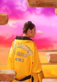 Kvinne i gul jakke med "UNLIMITED 24 HOURS KIKO" på ryggen, som står blant flytende steinplattformer med en livlig rosa og oransje himmel i bakgrunnen.