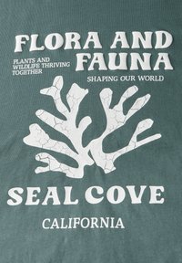 Zelené tričko s bílou, popraskanou typografií. Obsahuje korálovou grafiku a text: "FLORA A FAUNA," "SEAL COVE" a "CALIFORNIE."