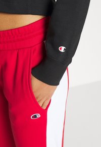 Persona che indossa pantaloni sportivi rossi e bianchi e una maglietta nera a maniche lunghe, con una mano appoggiata nella tasca dei pantaloni, visibile il logo Champion.