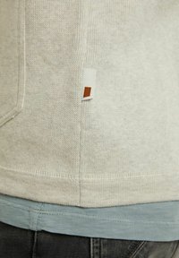 Gros plan de l'ourlet d'un pull beige tricoté avec une petite étiquette en tissu rectangulaire sur une chemise bleue et un pantalon foncé.