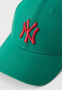 Grønn caps laget av stoff, med en fremtredende rød brodert "NY"-logo. Designet inkluderer en buet skjerm og søm detaljer.