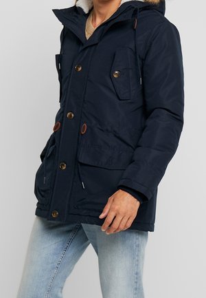 Veste matelassée bleu marine avec capuche en fourrure, dotée de plusieurs poches et d'accents en boutons marron, fabriquée dans un tissu lisse et durable.