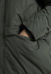 Chaqueta acolchada verde oscuro con una mano visible metiéndose en un bolsillo lateral. La tela tiene una textura suave y acentos de costura visibles.
