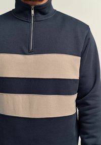 Navy sweatshirt with a high collar and quarter zip, featuring horizontal beige stripes across the chest and smooth fleece texture.