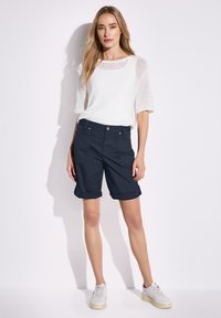 Top a maglia bianca con maniche corte, abbinato a pantaloni corti blu arrotolati. Il modello indossa sneakers bianche; look complessivo casual e rilassato.
