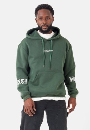 Grüne Kapuzenjacke mit weißem Aufdruck, ausgestattet mit einer Kängurutasche, verstellbarem Kordelzug an der Kapuze und gerippten Bündchen. Getragen über einem weißen T-Shirt und schwarzen Hosen.
